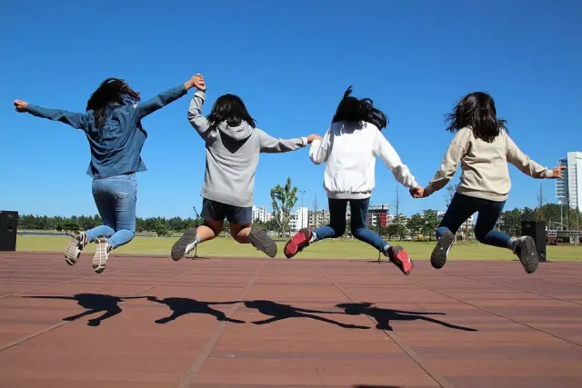 Na zdjęciu 4 dziewczęta trzymające się za ręce i wykonujące podskok nad szkolnym boiskiem. W tle błękitne niebo i widoczne z daleka bloki mieszkalne.