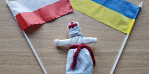 Na zdjęciu obok siebie flaga polski po lewej stronie po prawej flaga Ukrainy w środku tradycyjna ukraińska lalka motanka w bałym stroju przepasana haftowanym pasem.